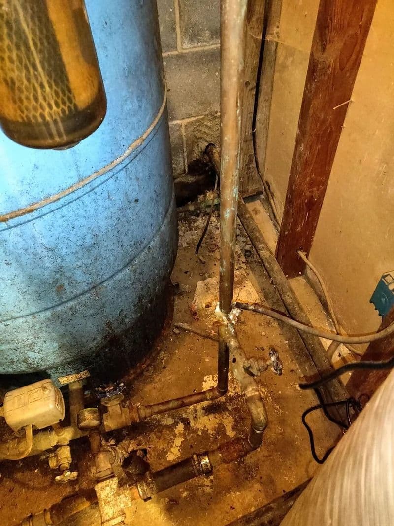 Blue tank with a pressure switch beside corroded copper and steel pipe fittings on a stained concrete floor