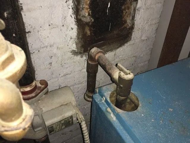 Rusted pipe elbow entering the top of a blue heating unit, below a soot-darkened opening in a painted brick wall