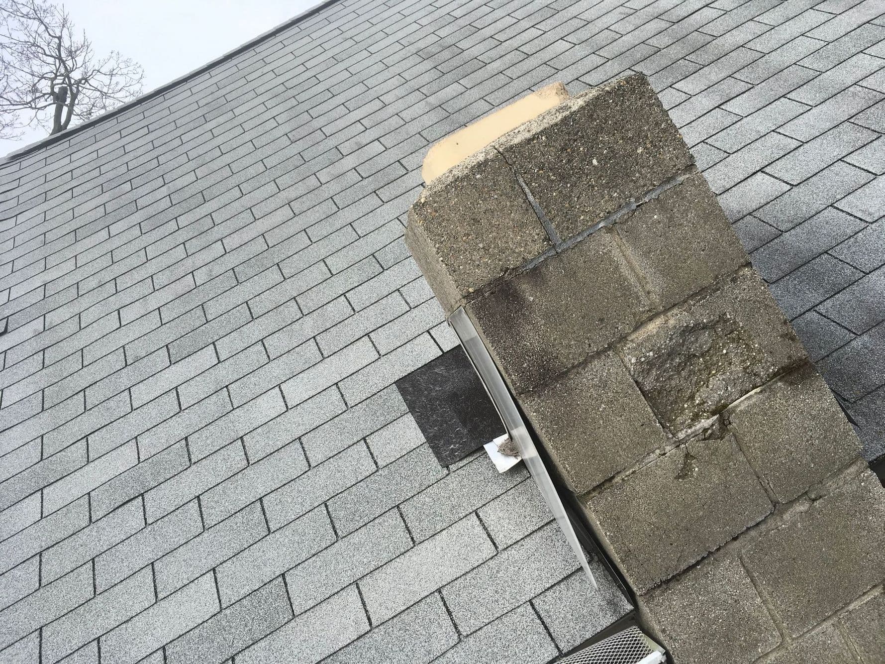 Concrete block chimney with a crumbling top and spalled block face, rising through an asphalt shingle roof