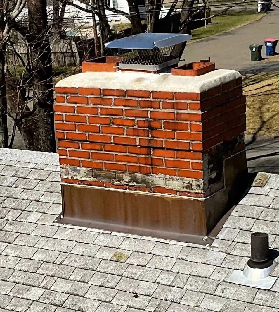 Brick chimney with a metal rain cap, worn and patched mortar near its base and brown metal flashing at the roof