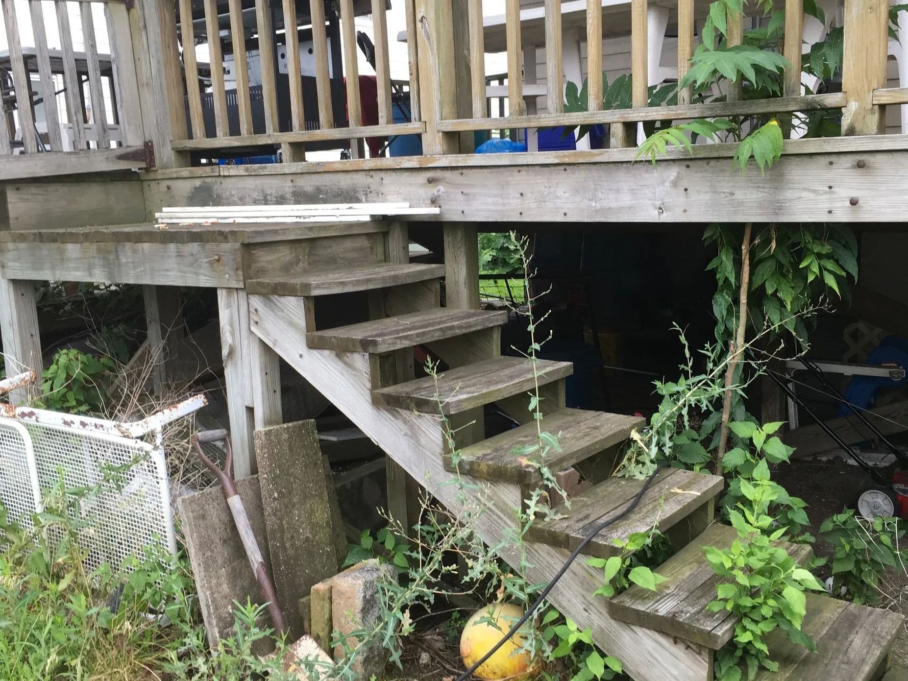 Weathered wooden deck stairs with worn treads, surrounded by overgrown weeds and loose concrete blocks