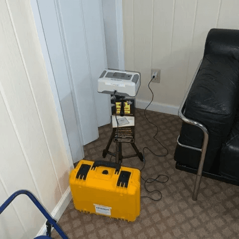Electronic radon monitor on a tripod, plugged into a wall outlet, next to a yellow hard carrying case