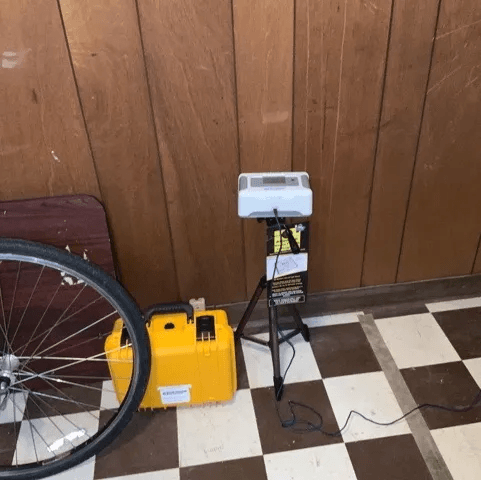 Radon monitor on a tripod against wood panelling on a checkerboard floor, with a yellow case and a bicycle wheel