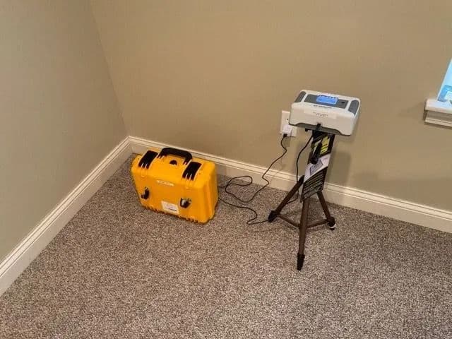 Radon monitor on a tripod in a carpeted room, plugged into an outlet, with a yellow hard case nearby