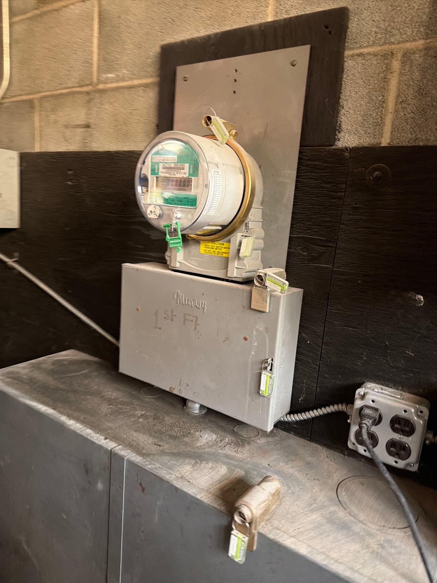 Electric meter with tamper seals on a grey enclosure marked 1st Fl, mounted on a board against a block wall