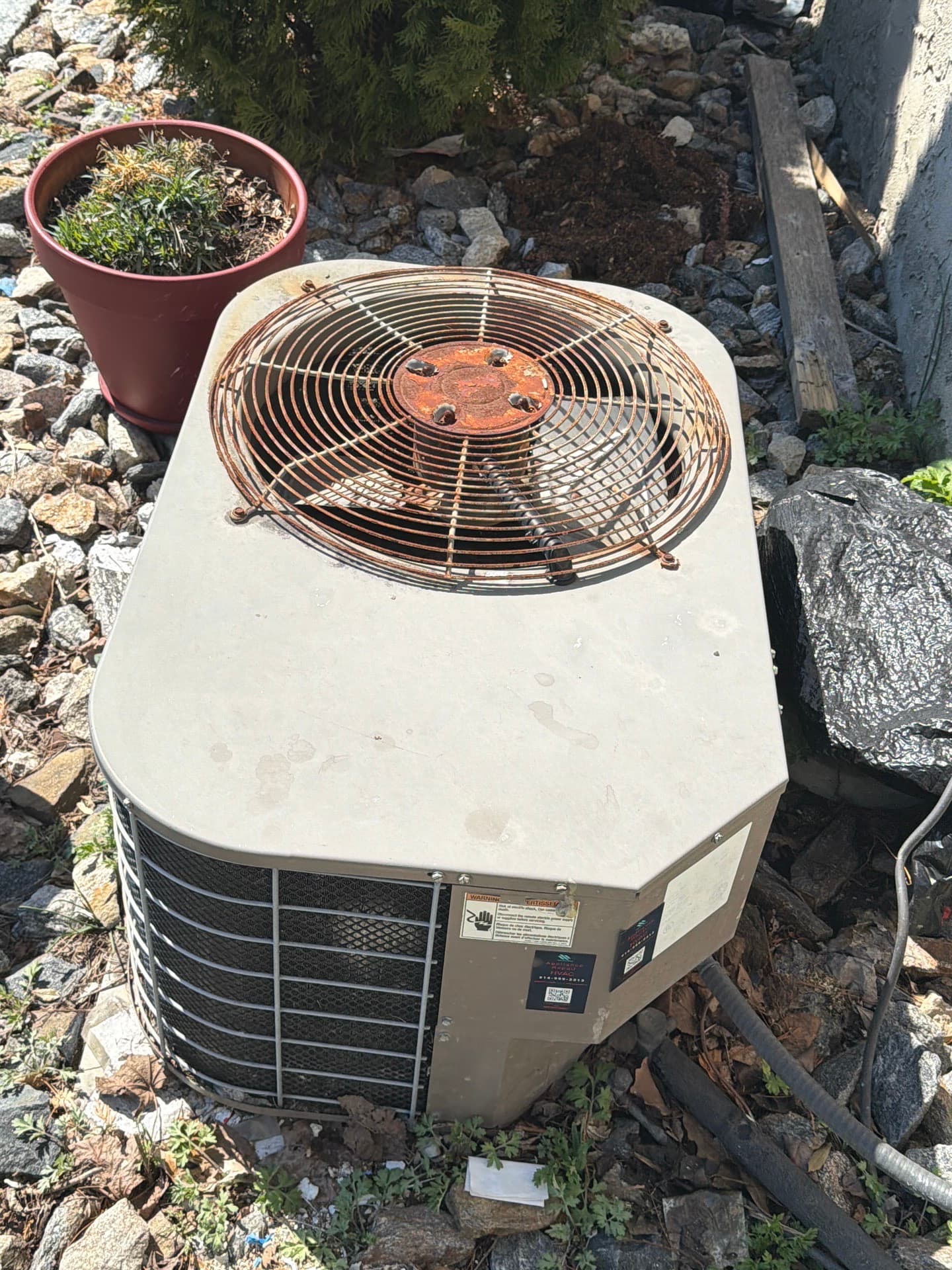 Outdoor air-conditioning condenser with a rusted fan guard and motor cap, set among rocks and debris