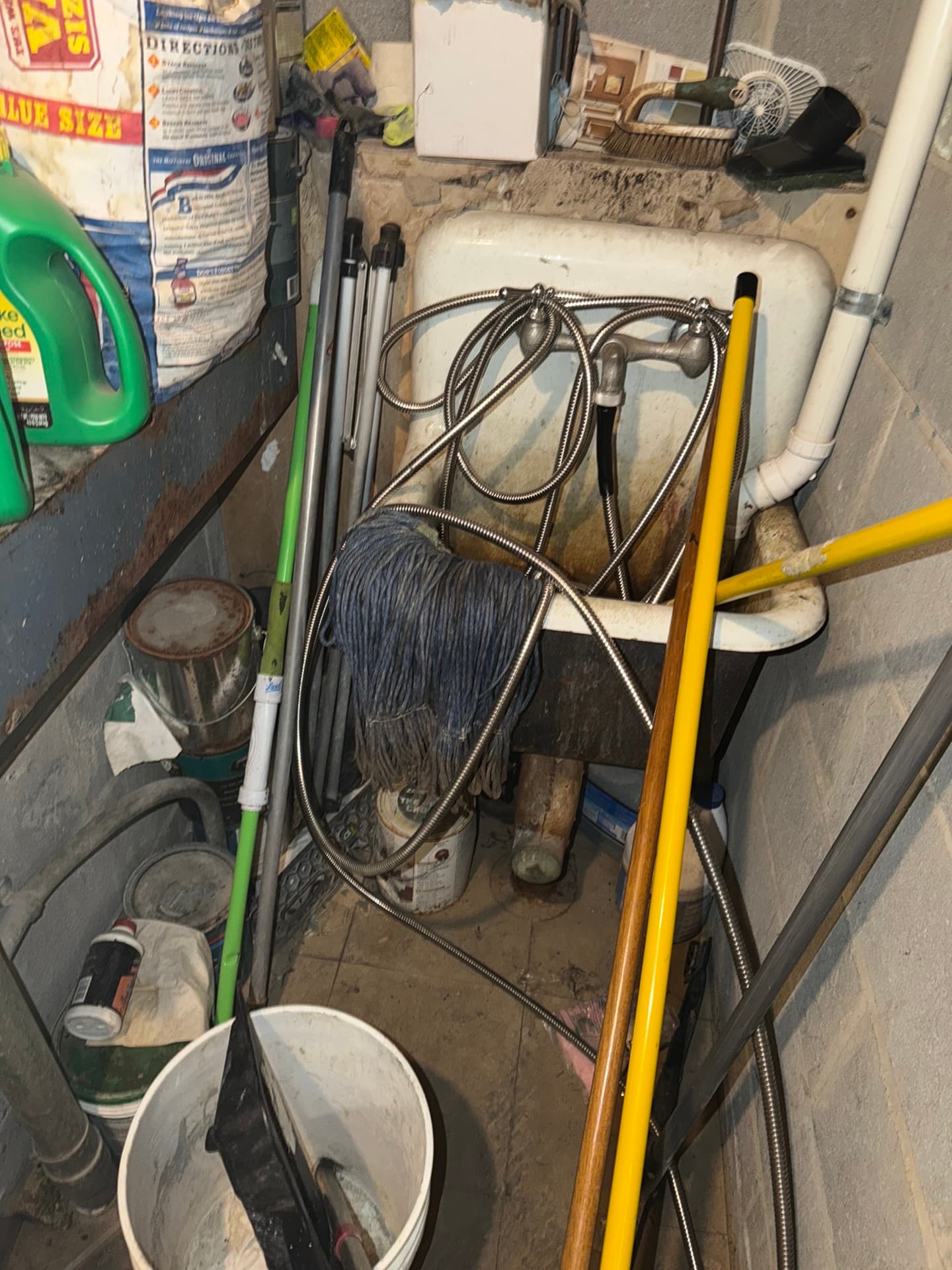 Utility sink crowded with mops, brooms, braided hoses, buckets and paint cans in a corner with block walls